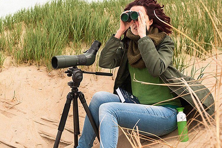 Een persoon in de duinen met een telescoop op een statief en een verrekijker in de hand, op zoek naar vogels